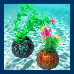 May include: Two decorative ceramic pots with green and pink artificial plants. The pots are round and have a metallic finish. The pot on the left is blue and the pot on the right is copper.
