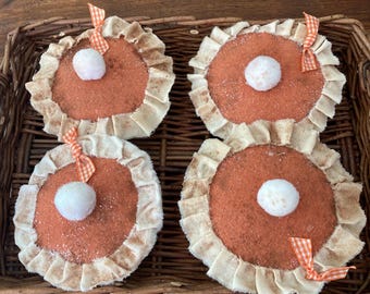 May include: Four decorative, round, felt pie ornaments with orange-brown centers, cream-colored ruffled edges, and white pom-pom centers. Each pie has a small orange and white checkered ribbon bow. The pies are arranged in a woven basket.