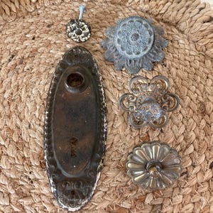 May include: Collection of antique door hardware, including ornate knobs and a keyhole plate. The knobs feature various designs, including floral and geometric patterns, with finishes in silver, bronze, and distressed white. The hardware is arranged on a woven mat.