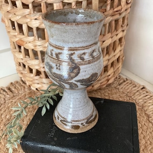 May include: A handmade, ceramic goblet with a wide rim and pedestal base. The goblet is light gray with brown decorative patterns around the body. It rests on a black book, with a woven basket in the background.