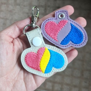 May include: Two heart-shaped keychains with embroidered designs. One keychain is purple with a pink heart and a blue heart. The other keychain is white with a pink, yellow, and blue heart.