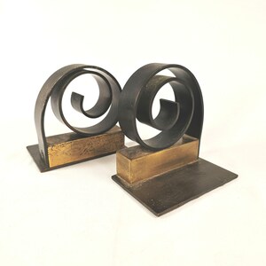 May include: Pair of metal bookends with a dark, matte finish. Each bookend features a rectangular base with a gold-colored accent and a swirling, spiral design. The bookends are designed to hold books upright.