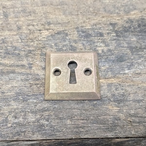 May include: A square-shaped keyhole cover made of metal with a brushed finish. The cover has a keyhole in the center and two small holes on either side. The cover is mounted on a piece of weathered wood.