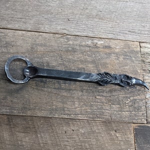 May include: Hand-forged metal bottle opener with a circular opening and a decorative bird head design. The opener is made of dark metal and has a rustic, handcrafted appearance. The bird head is detailed with feathers.