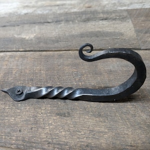 May include: A black metal hook with a twisted stem and a decorative curl at the top. The hook is mounted to a wooden surface with a screw.