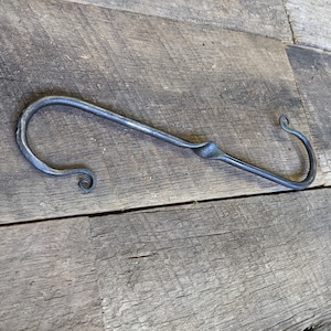 May include: A hand-forged metal hook with a twisted design and a decorative curl at each end. The hook is made of dark metal and is lying on a wooden surface.
