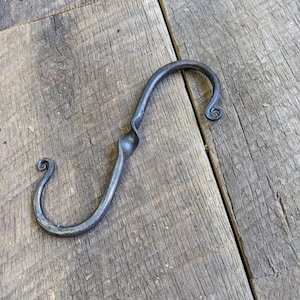 May include: A hand-forged metal hook with a unique S-shape and a curled end. The hook is made of dark metal and has a rustic, handmade look.