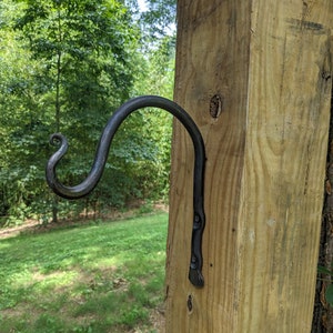 May include: A hand-forged, black metal hook mounted on a wooden post. The hook has a decorative, curled end and is attached to the post with rivets. The wood has a natural, light brown color.