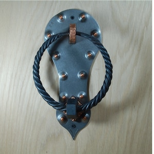 May include: A silver door knocker with a black rope ring. The knocker has a decorative design with copper accents and rivets.