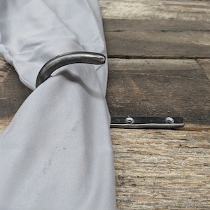May include: A black metal hook with a curved arm, mounted on a wooden surface. A gray fabric is draped over the hook.
