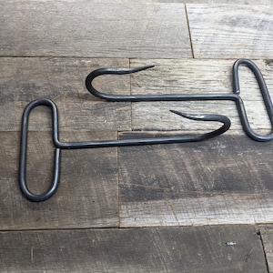 May include: Two black metal meat hooks with a T-shaped handle. The hooks are lying on a wooden surface.