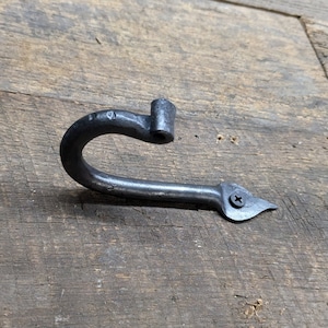 May include: A black metal hook with a curved design and a pointed end. The hook is attached to a wooden surface with a screw.