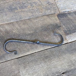 May include: A black metal hook with a twisted design and curled ends. The hook is lying on a wooden surface.