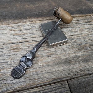 May include: A handcrafted, metal cigar holder with a skull design at the handle's end, resting on a silver lighter. The cigar is lit and held in place by the holder. The items are on a wooden surface.