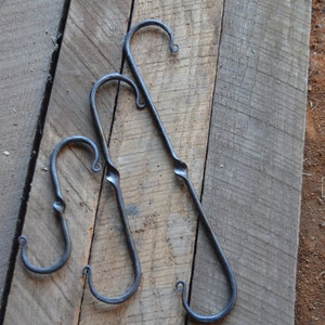 May include: Three black metal hooks of different sizes, hanging on a wooden surface. The hooks are all curved and have a twisted design.