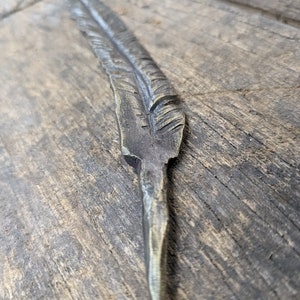 May include: A metal feather with a detailed design, crafted in a rustic style. The feather is a decorative piece with a pointed tip and a textured surface.