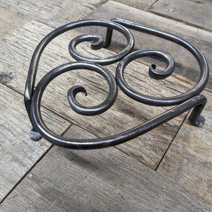 May include: Hand-forged metal trivet with a decorative, swirling design. The trivet is made of dark gray metal and features three curled spirals and three small feet for stability. A rustic, handcrafted kitchen accessory.