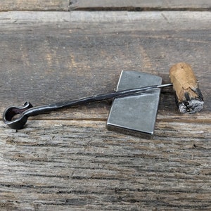 May include: A silver lighter with a black metal rod attached to a metal loop. The lighter is sitting on a wooden surface next to a brown and black cigar.