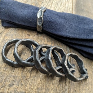 May include: Five black metal napkin rings, with a hammered finish, are stacked on a wooden surface. The rings are designed to hold napkins in place at a dinner table.