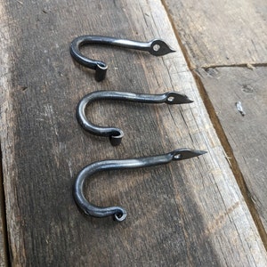 May include: Three hand-forged metal hooks with a dark, textured finish. Each hook has a curved body and a pointed end with a small hole. The hooks are arranged on a weathered wooden surface, showcasing their rustic aesthetic.