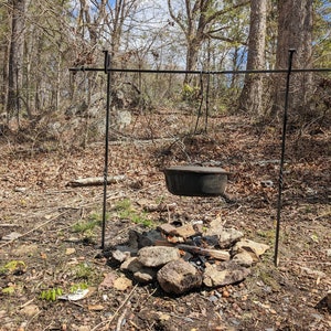 May include: A black cast iron pot hanging over a campfire on a tripod stand in a wooded area.