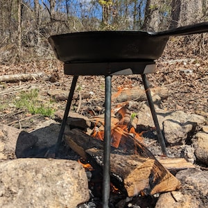 May include: A black cast iron skillet sits on a metal tripod over a small campfire. The tripod has three legs and is made of sturdy metal. The campfire is made of logs and is burning brightly.