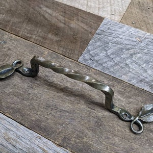 May include: A twisted metal door handle with leaf-shaped accents. The handle is a dark bronze color and has a rustic look.