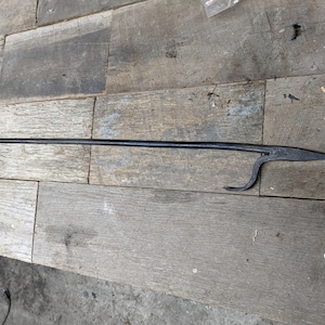 May include: A black, wrought iron fireplace poker with a looped handle and a curved hook at the end. The poker is long and slender, with a pointed tip opposite the handle. The tool rests on a weathered wooden surface.