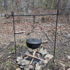 May include: A black metal tripod stand with a hanging pot over a campfire. The pot has a lid and is hanging from a hook on the stand.