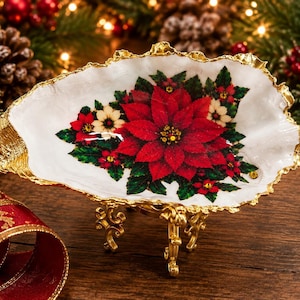 Peut inclure: Un plat décoratif en forme de coquille d'huître orné d'un motif de poinsettia rouge vif et de feuilles de houx vertes sur un fond blanc. La coquille présente un bord texturé peint en doré et repose sur un petit support en métal doré à quatre pieds.
