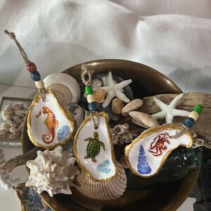 May include: Three painted oyster shell ornaments with sea creature designs, including a seahorse, turtle, and crab. Each shell has a gold painted edge and is strung with a beaded cord. The shells are displayed in a brass bowl with seashells and starfish.
