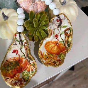 May include: Two decorative oyster shell ornaments featuring pumpkin and autumn leaf designs, with gold trim. Each shell is strung with white beads. The background includes small pumpkins and a succulent plant, creating an autumnal theme.