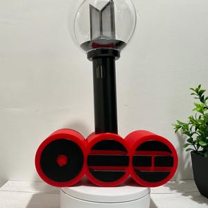 May include: A black and clear spherical light stick with a BTS logo inside, mounted on a red three-part circular stand. The stand features black horizontal and circular cutouts. The light stick has a long black handle and a transparent globe top.
