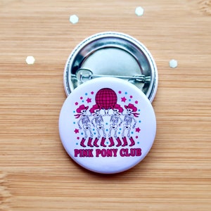May include: White button with a pink disco ball and five skeletons wearing cowboy hats and boots. The text "PINK PONY CLUB" is printed below the skeletons.