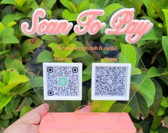 May include: A pink and white sign with the text "Scan To Pay" in a stylized font. The sign also includes the text "We also accept cash & cards!" and has two QR codes, one for Cash App and one for Venmo. The sign is on a pink base with the text "the little kumquat" in white.