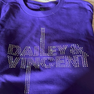 May include: A purple t-shirt with the text "DAILEY & VINCENT" spelled out in sparkling rhinestones. The text is centered on the front of the shirt. The shirt is folded.