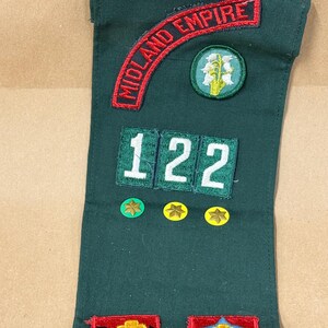 May include: A dark green sash with embroidered patches. The patches include the words "MIDLAND EMPIRE" in red, numbers "122", and various star and floral designs. The sash is likely for a youth organization.