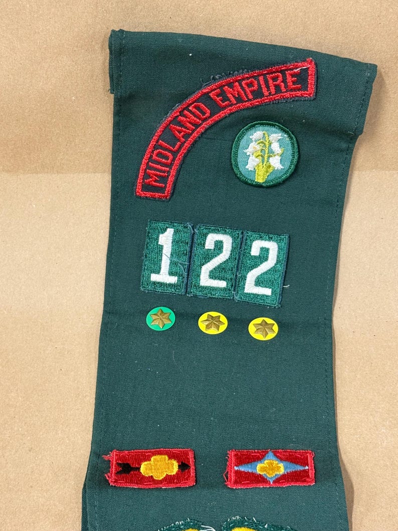 May include: A dark green sash with embroidered patches. The patches include the words "MIDLAND EMPIRE" in red, numbers "122", and various star and floral designs. The sash is likely for a youth organization.