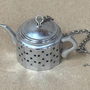 May include: A silver-colored metal tea infuser shaped like a miniature teapot. The body features a series of small circular perforations for steeping. It includes a removable lid with a small loop and a metal chain with a hook attachment.
