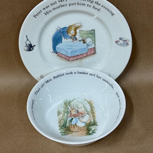 May include: A white ceramic plate and bowl set featuring Peter Rabbit illustrations. The plate shows a rabbit in bed with the text, "Peter was not very well during the evening. His mother put him to bed." The bowl depicts a rabbit with a basket.