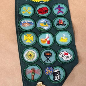 May include: A dark green sash with multiple embroidered patches. The patches feature various designs, including a yellow cross, a campfire, and a lighthouse. A gold-colored emblem with wings is at the top.