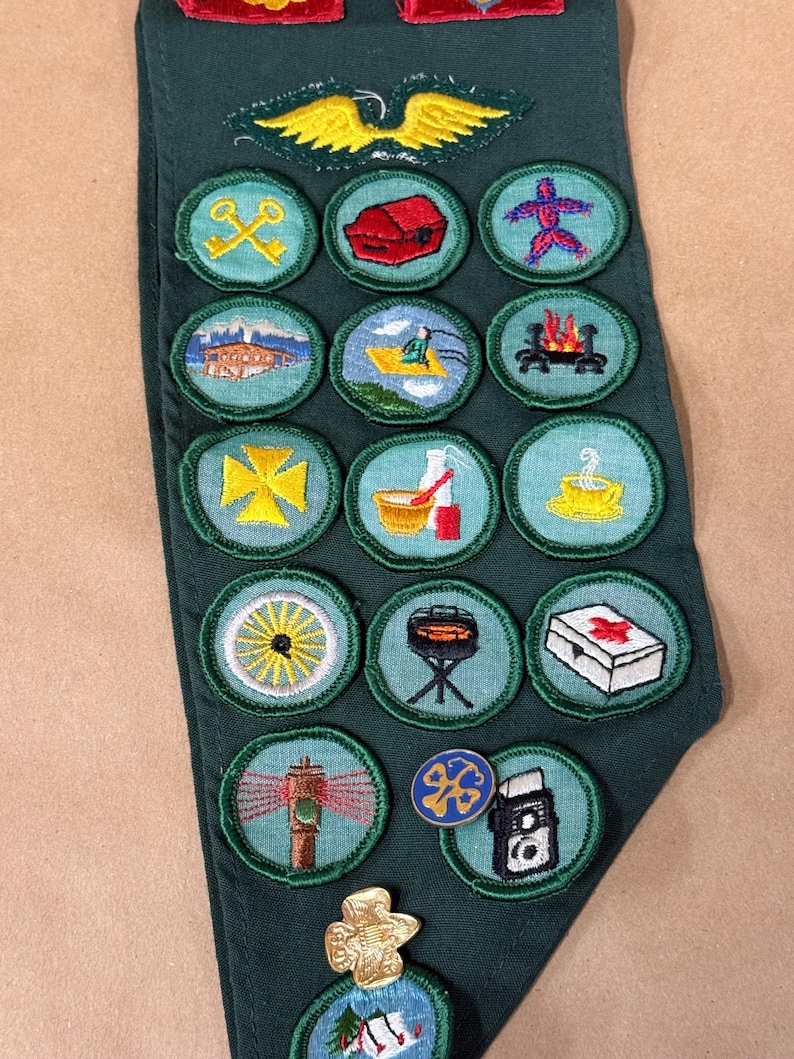 May include: A dark green sash with multiple embroidered patches. The patches feature various designs, including a yellow cross, a campfire, and a lighthouse. A gold-colored emblem with wings is at the top.
