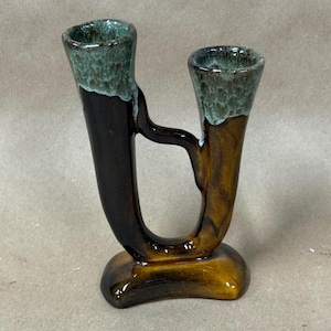 May include: A ceramic vase with two openings, featuring a unique, curved design. The vase is primarily brown with a glossy finish, transitioning to a darker brown and topped with a green, textured rim. The base is rounded.