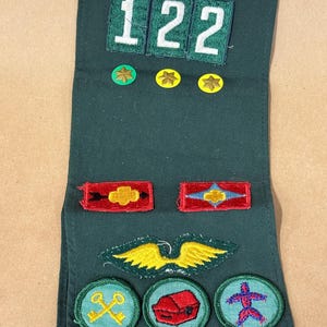 May include: Dark green sash with various embroidered patches. Patches include the number "122", stars, and circular designs with images of a building, a treasure chest, and a person. The sash is likely for a youth organization.