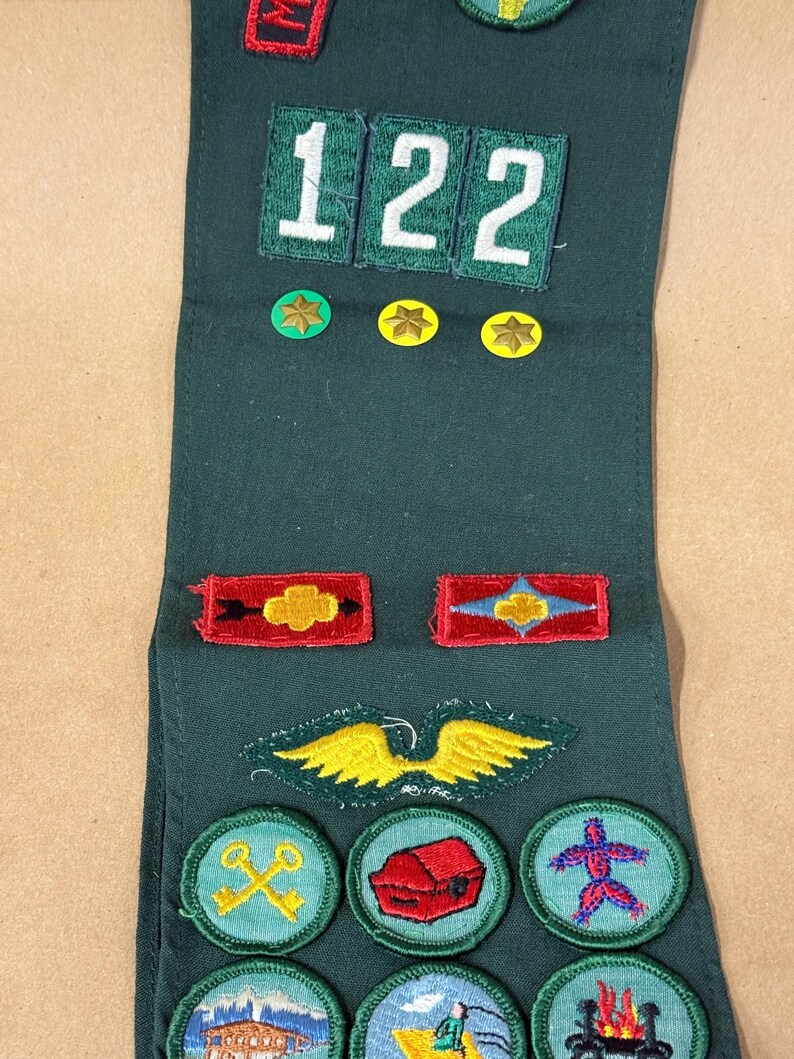 May include: Dark green sash with various embroidered patches. Patches include the number "122", stars, and circular designs with images of a building, a treasure chest, and a person. The sash is likely for a youth organization.