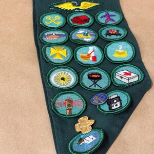 May include: A dark green neckerchief with various embroidered patches and pins. The patches feature images of a yellow eagle, a campfire, a lighthouse, and other symbols. The neckerchief is made of a soft fabric.