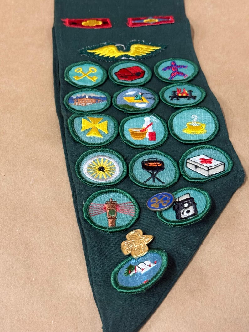 May include: A dark green neckerchief with various embroidered patches and pins. The patches feature images of a yellow eagle, a campfire, a lighthouse, and other symbols. The neckerchief is made of a soft fabric.