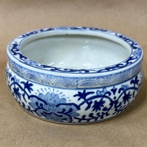 May include: A round ceramic bowl featuring a white base with blue floral patterns and intricate scrollwork. The exterior displays a repeating design of stylized flowers and vines, while the rim is decorated with a thin band of wave-like motifs.