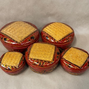 May include: Five red, round decorative boxes with woven, light brown lids. The boxes have a glossy finish and are adorned with floral patterns in gold and black. The lids are square-shaped and the boxes are of varying sizes.