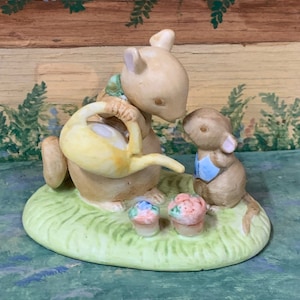 May include: A ceramic figurine featuring two mice and two small flower pots on a green base. One mouse holds a yellow watering can, while the other mouse wears a blue vest. The figurine has a light brown color.